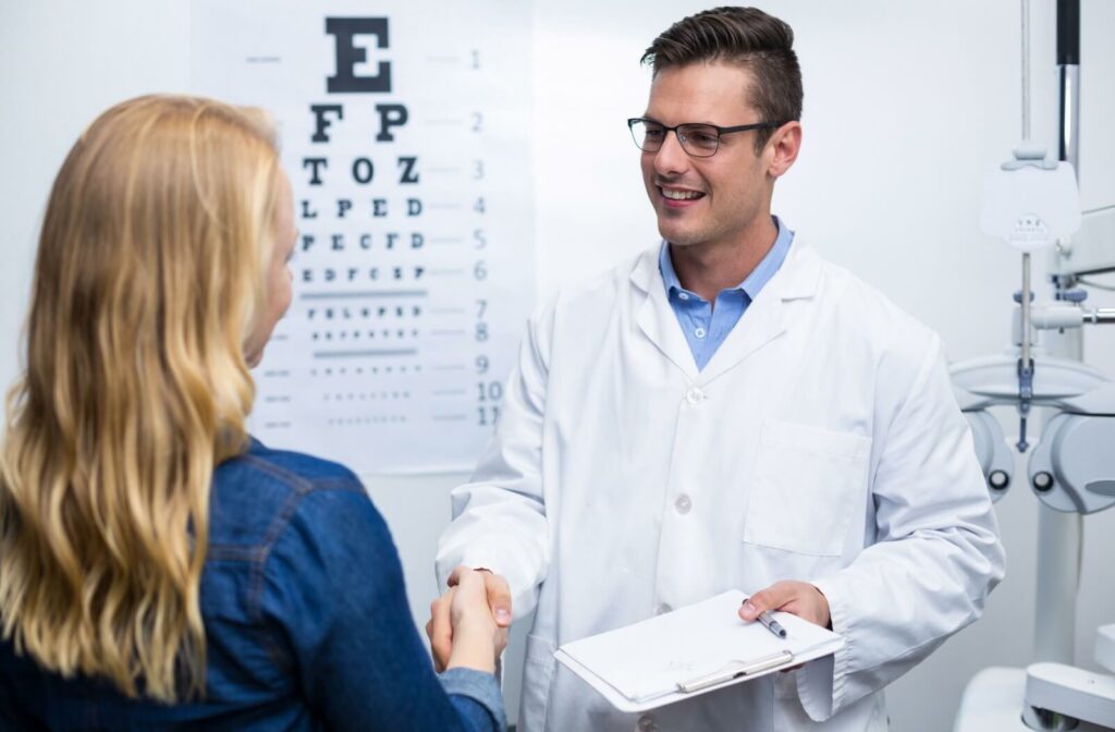 A patient with long blond hair shakes hands with their optometrist, who is smiling and holding a clipboard.