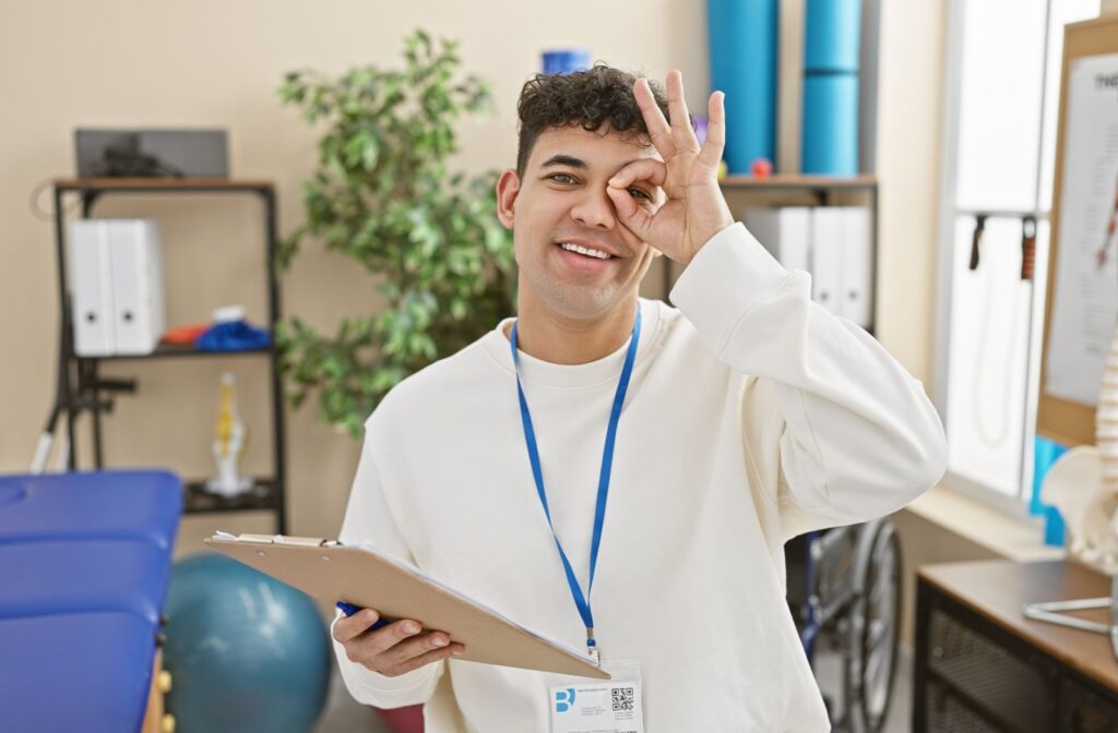 A vision therapist in a bright, welcoming office smiles and holds up an OK sign by their eye to indicate good visual health