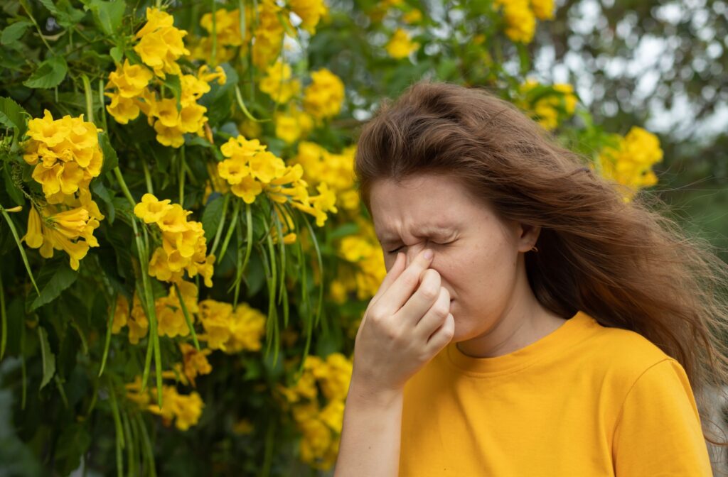 A young woman wearing a yellow shirt rubbing her eyes, which are irritated by pollen from the yellow flowers around her.