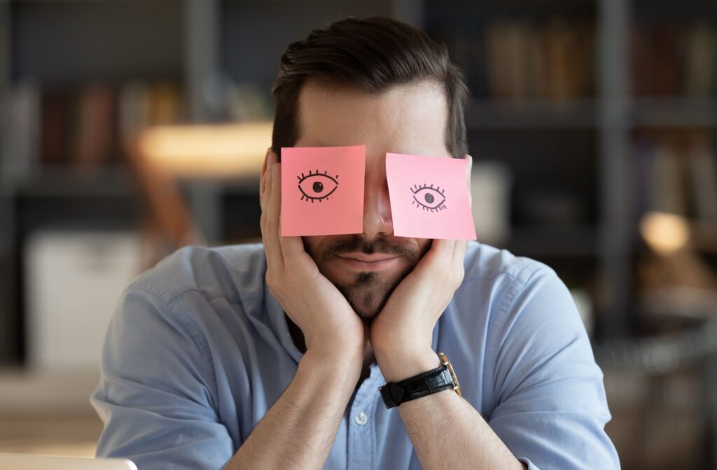 A man rests his head in his hands with two sticky notes over his eyes. Cartoon eyes are drawn on the sticky notes, representing nocturnal lagophthalmos.