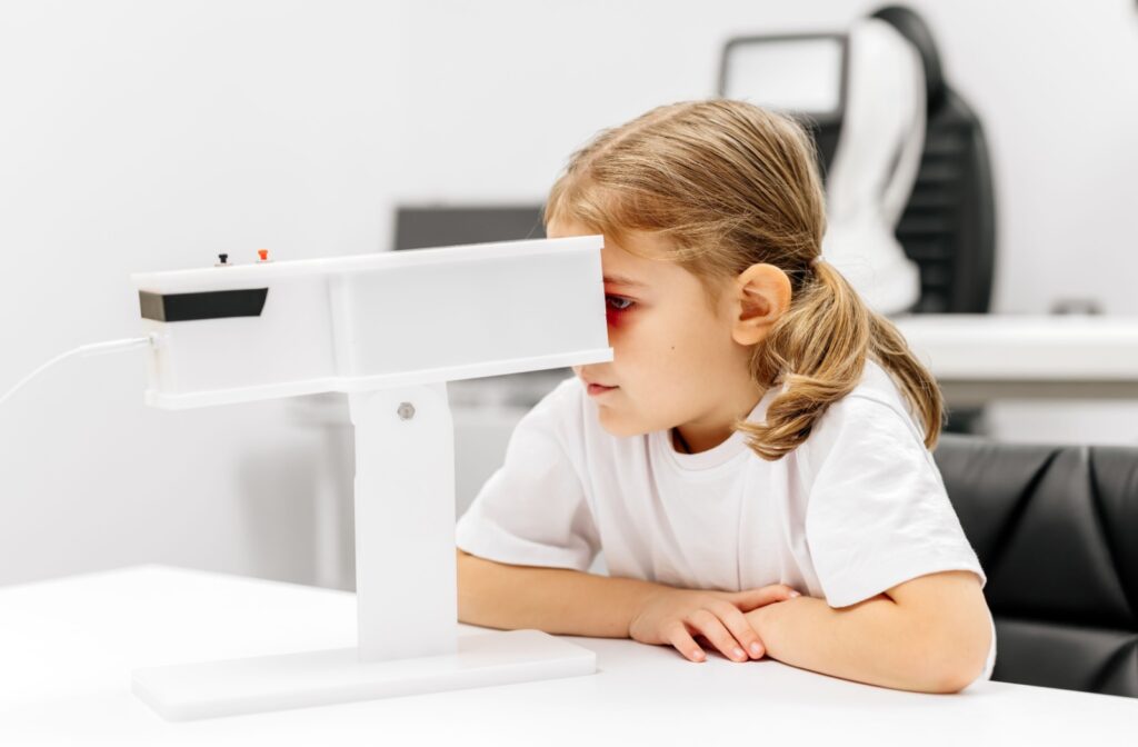 A child sits at a white table during an eye exam, looking into a diagnostic vision screening device.