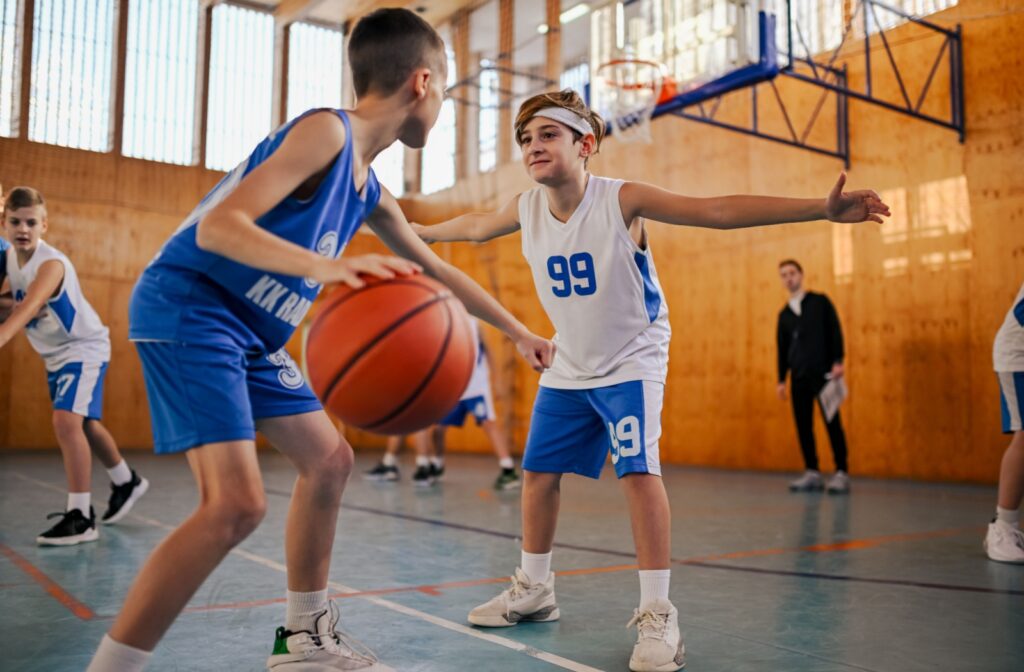 Two children in basketball uniforms play against each other on an indoor court.