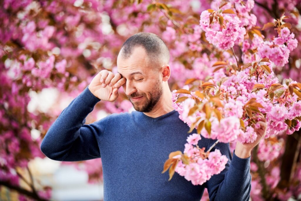 A person standing in front of a pink blossoming tree and rubbing their irritated eyes